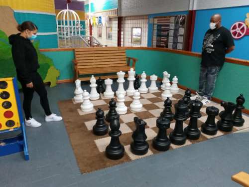 Two people in masks play a large outdoor chess game in a colorful indoor space.