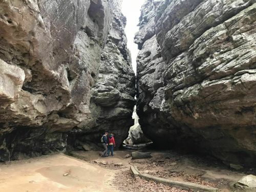 Two people stand between towering rock formations in a narrow canyon, with a small waterfall visible above.