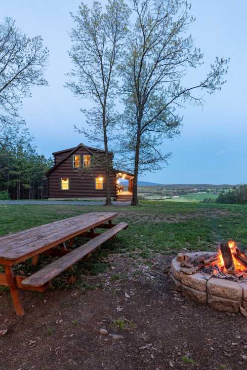 A cozy cabin at dusk, with a picnic table and a fire pit in the foreground, surrounded by trees and open fields.