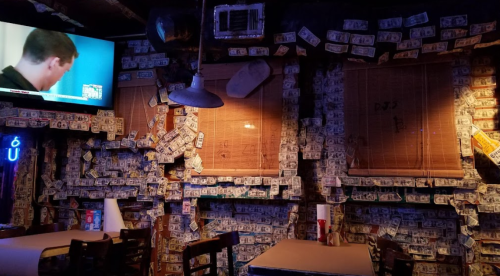 A dimly lit bar with walls covered in dollar bills and a TV showing a program in the background.