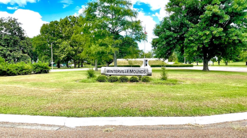 Sign for Winterville Mounds surrounded by green grass and trees under a blue sky with scattered clouds.
