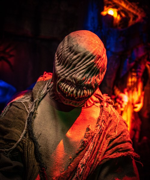 A creepy figure with a distorted, toothy face, wearing tattered clothing, set against a dark, colorful background.
