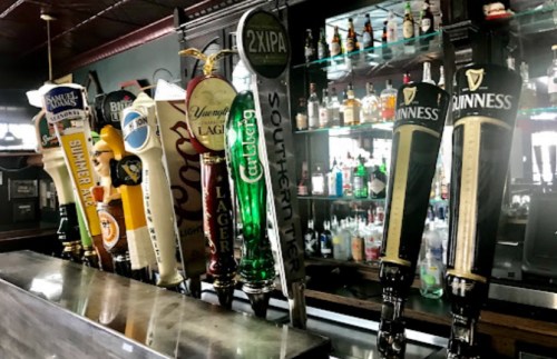 A row of beer taps featuring various brands, with a bar shelf of bottles in the background.