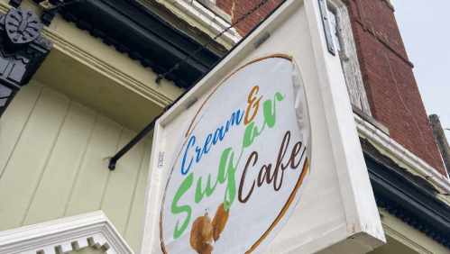 Sign for "Cream & Sugar Cafe" featuring colorful lettering on a white background, hanging from a building.