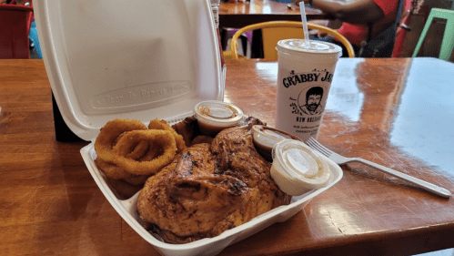 A takeout container with grilled chicken, onion rings, dipping sauces, and a drink on a wooden table.