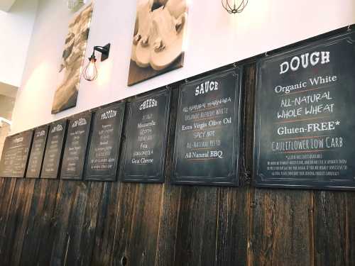 Menu boards on a wooden wall display options for dough, cheese, sauce, and protein in a casual dining setting.