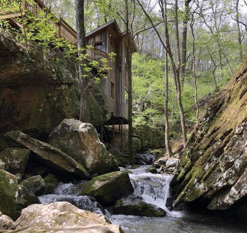 A wooden cabin nestled among rocks and trees beside a flowing stream in a lush, green forest.