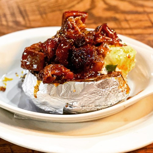 A baked potato topped with savory meat and sauce, served on a white plate with a foil wrapper.
