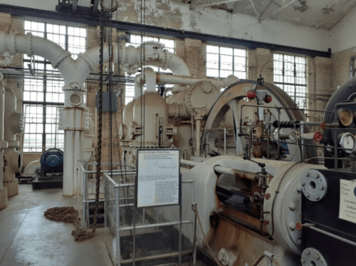 Interior of an industrial facility featuring large machinery, pipes, and windows, showcasing vintage equipment.