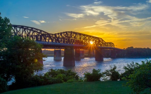 A sunset casts golden light through a bridge over a river, surrounded by trees and a clear sky.