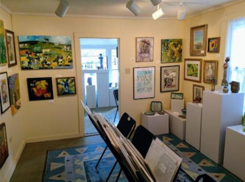 Art gallery interior featuring various paintings and sculptures displayed on walls and pedestals, with chairs in the foreground.