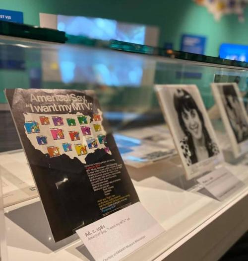 A display featuring a vintage "I Want My MTV" advertisement and black-and-white photos in a museum setting.