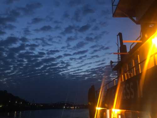 A boat illuminated by warm lights against a twilight sky filled with clouds, reflecting on calm water.