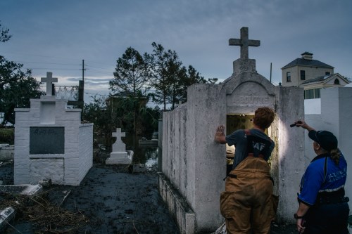A person shines a flashlight into a crypt in a cemetery, surrounded by tombstones and a cloudy sky.
