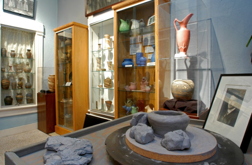 A pottery display in a museum featuring various ceramic pieces and a pottery wheel with clay and rocks.