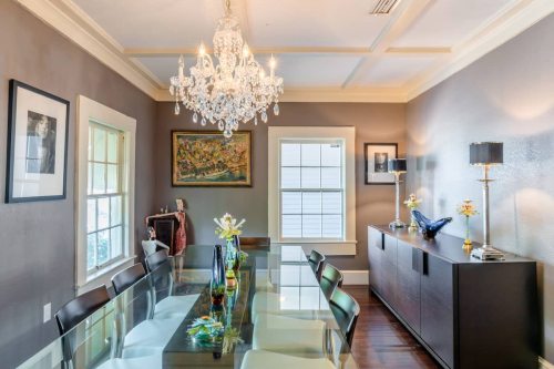 A modern dining room featuring a glass table, black chairs, a chandelier, and elegant decor on the walls and sideboard.
