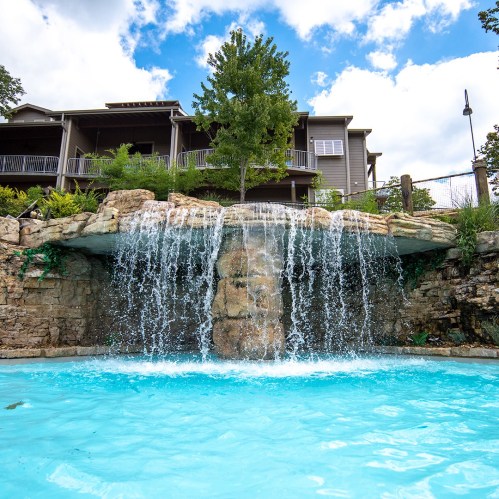A serene pool with a waterfall, surrounded by lush greenery and a modern building in the background under a blue sky.
