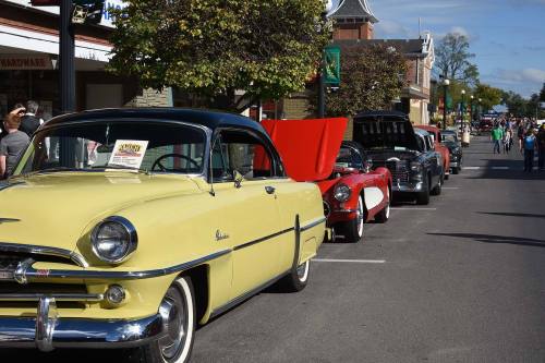 A row of classic cars parked on a street, with a yellow car in the foreground and people in the background.