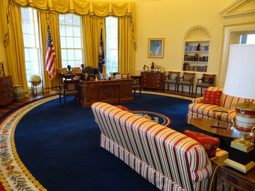 The Oval Office featuring a large desk, striped sofas, a globe, and yellow curtains with the American flag.