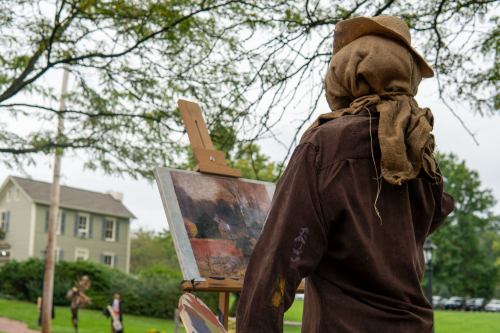 A scarecrow stands before an easel, painting a landscape, with trees and a house in the background.