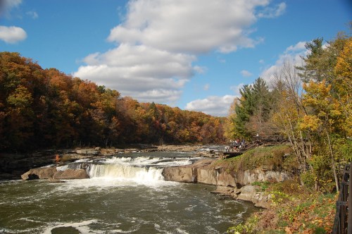 A scenic view of a waterfall surrounded by colorful autumn foliage under a partly cloudy sky.