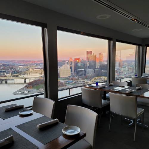 A dining area with modern tables and chairs, featuring a panoramic view of a city skyline at sunset.