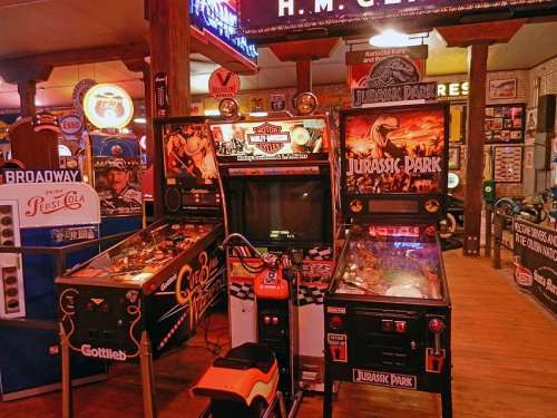 Two vintage arcade machines, one for "Jurassic Park" and another for "Harley Davidson," in a retro game room setting.