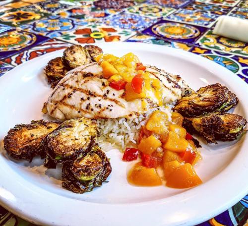 Grilled fish on rice, surrounded by roasted Brussels sprouts and topped with mango salsa, on a colorful tiled table.