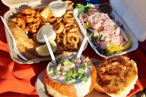 A variety of food including curly fries, a salad, and a seafood chowder in a bread bowl, served in takeout containers.