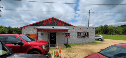 A BBQ restaurant named "Hank's BBQ" with a gray exterior and red accents, surrounded by parked cars and a grassy area.