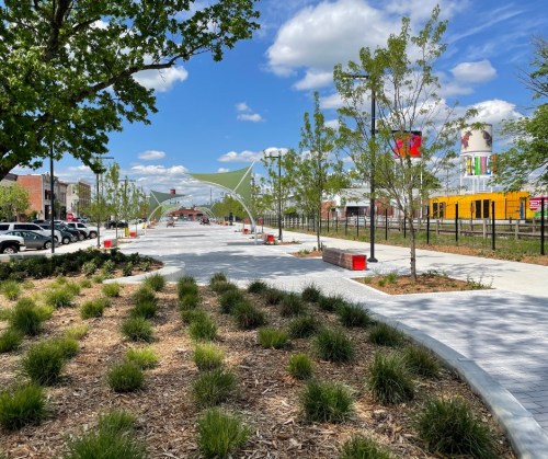 A sunny park pathway lined with greenery, benches, and colorful banners, leading to a vibrant urban area.