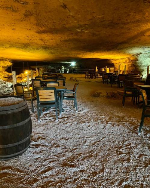 A dimly lit cave interior with wooden tables and chairs, surrounded by stone walls and a barrel in the foreground.