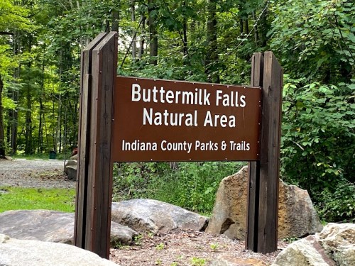 Sign for Buttermilk Falls Natural Area, surrounded by trees and rocks, indicating Indiana County Parks & Trails.