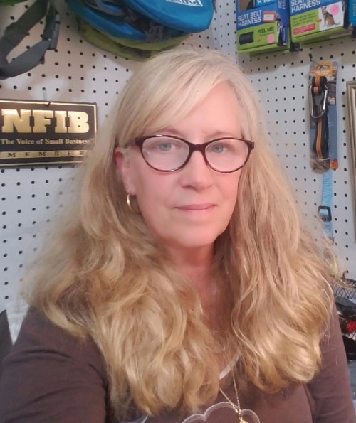 A woman with long, wavy blonde hair and glasses smiles at the camera, surrounded by tools and equipment on a pegboard.