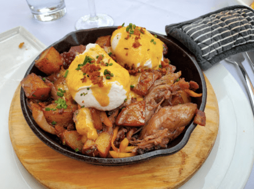 A skillet dish with poached eggs, hollandaise sauce, crispy potatoes, and pulled meat, garnished with chives.