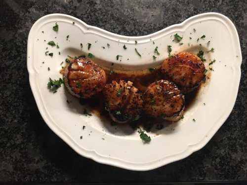 A white plate with four seared scallops garnished with herbs and a dark sauce, set on a dark countertop.