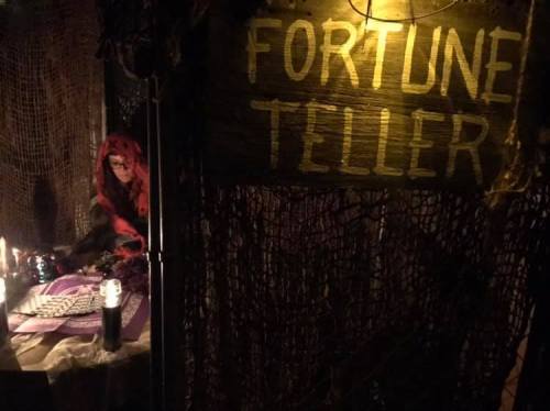 A dimly lit fortune teller booth with a sign, candles, and a person in a red cloak sitting at a table.