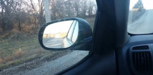 A car's side mirror reflects a rural road and trees in the background, with a clear sky above.