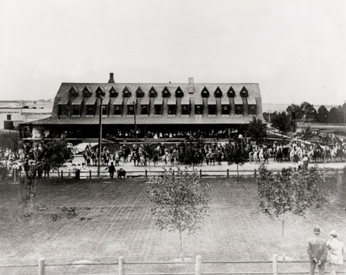 Historic black-and-white photo of a large building with a crowd and horses outside, surrounded by trees and a fence.