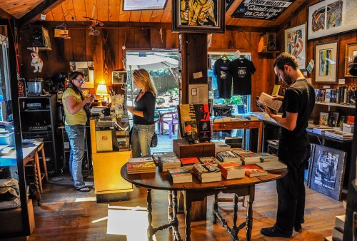 A cozy bookstore interior with wooden decor, featuring two customers browsing and a man reading at a table.