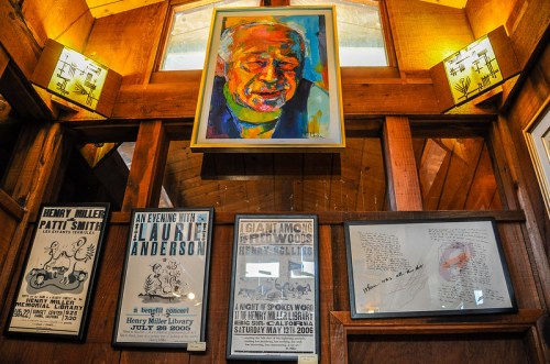 A wooden wall displays framed art and posters, including a colorful portrait and event announcements for Henry Miller.