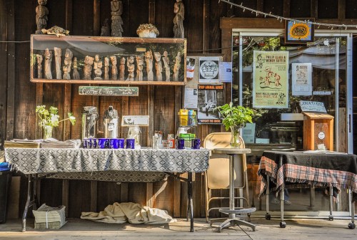 A rustic outdoor café setup with a table, coffee supplies, and decorative wooden sculptures against a wooden wall.