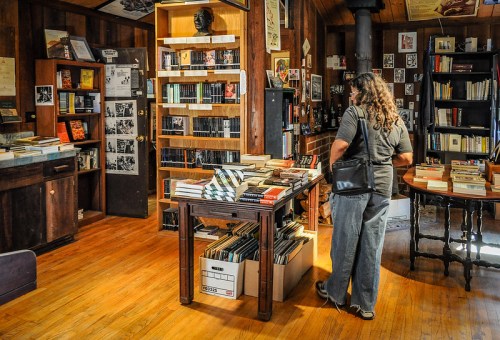 A person browses books in a cozy, wooden bookstore filled with shelves and a table of stacked books.