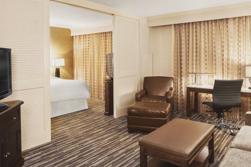 A hotel room featuring a sitting area with a chair, ottoman, desk, and a view of a bedroom through sliding doors.