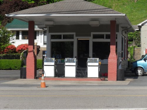 A small gas station with three fuel pumps and a service kiosk, surrounded by greenery and residential buildings.