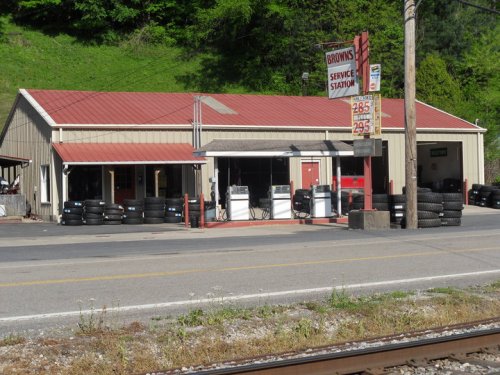 A service station with a red roof, gas pumps, and tires displayed outside, set along a rural road.