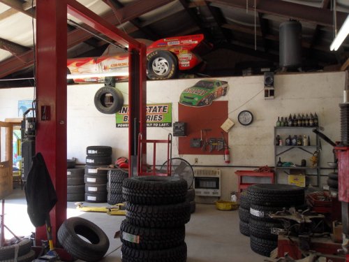 A garage with stacked tires, tools, and a race car hanging from the ceiling, featuring a racing-themed wall decoration.