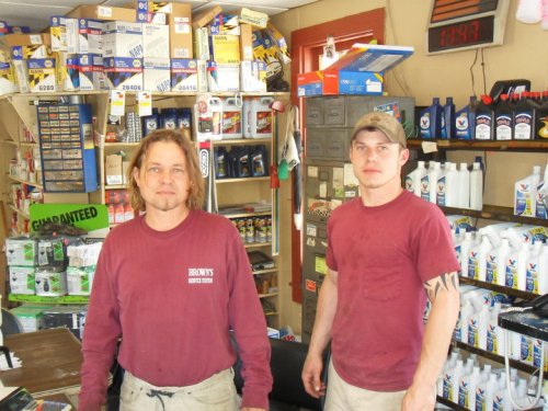 Two men in maroon shirts stand in a shop filled with automotive supplies and products. Shelves are stocked behind them.