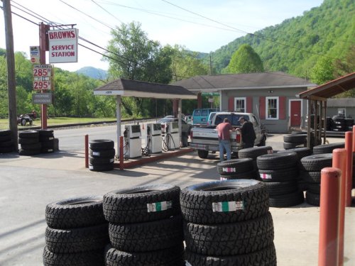 A service station with tire stacks, a gas pump, and a customer filling up a truck in a rural setting.
