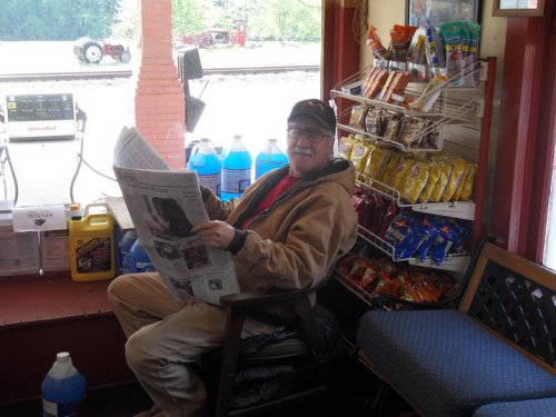 A man in a brown jacket sits in a chair, reading a newspaper, surrounded by snacks and drinks in a small store.